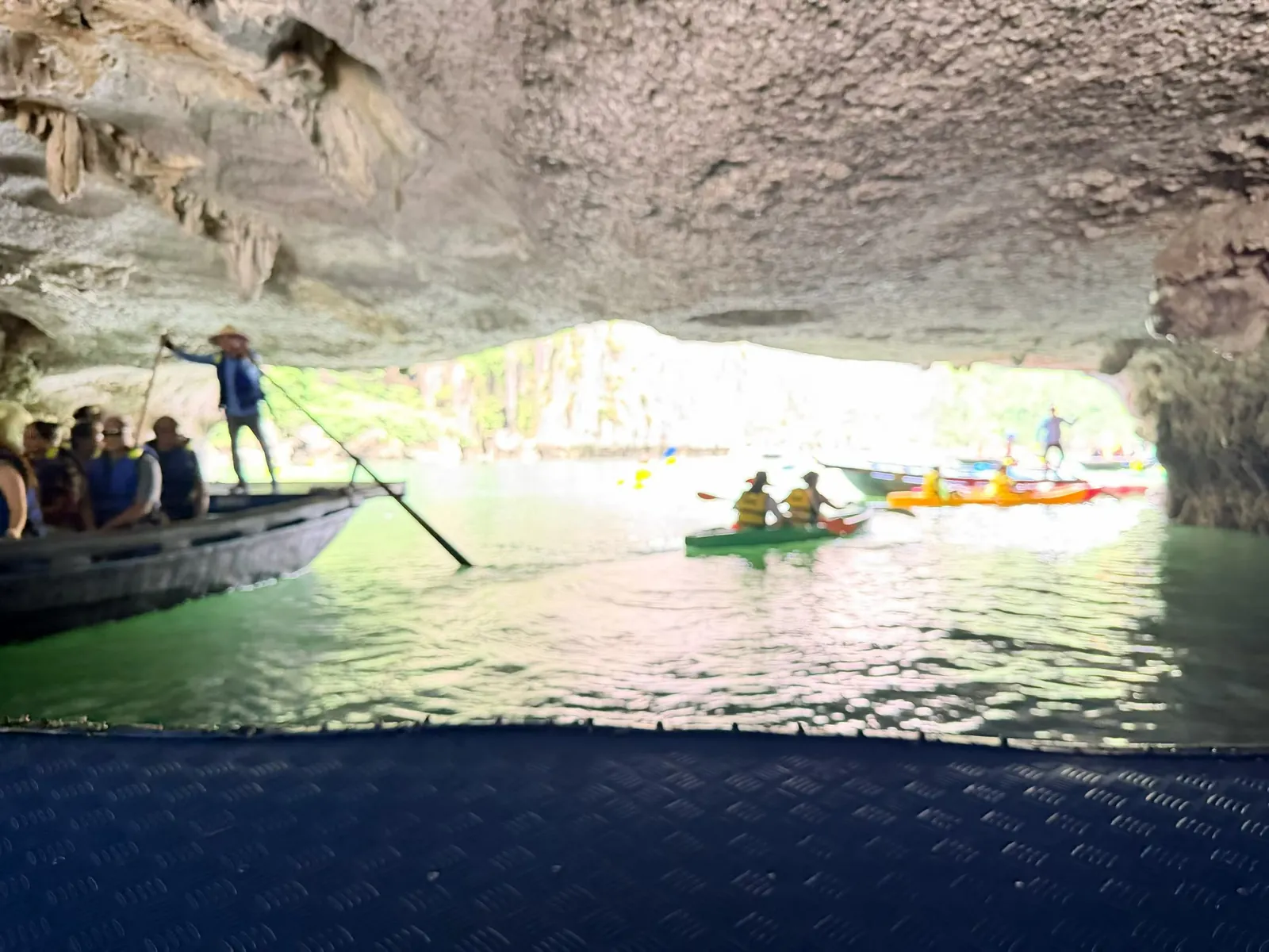 Inside a natural cave, a boatman poles a bamboo raft of visitors while kayaks float on the water beyond the cave mouth