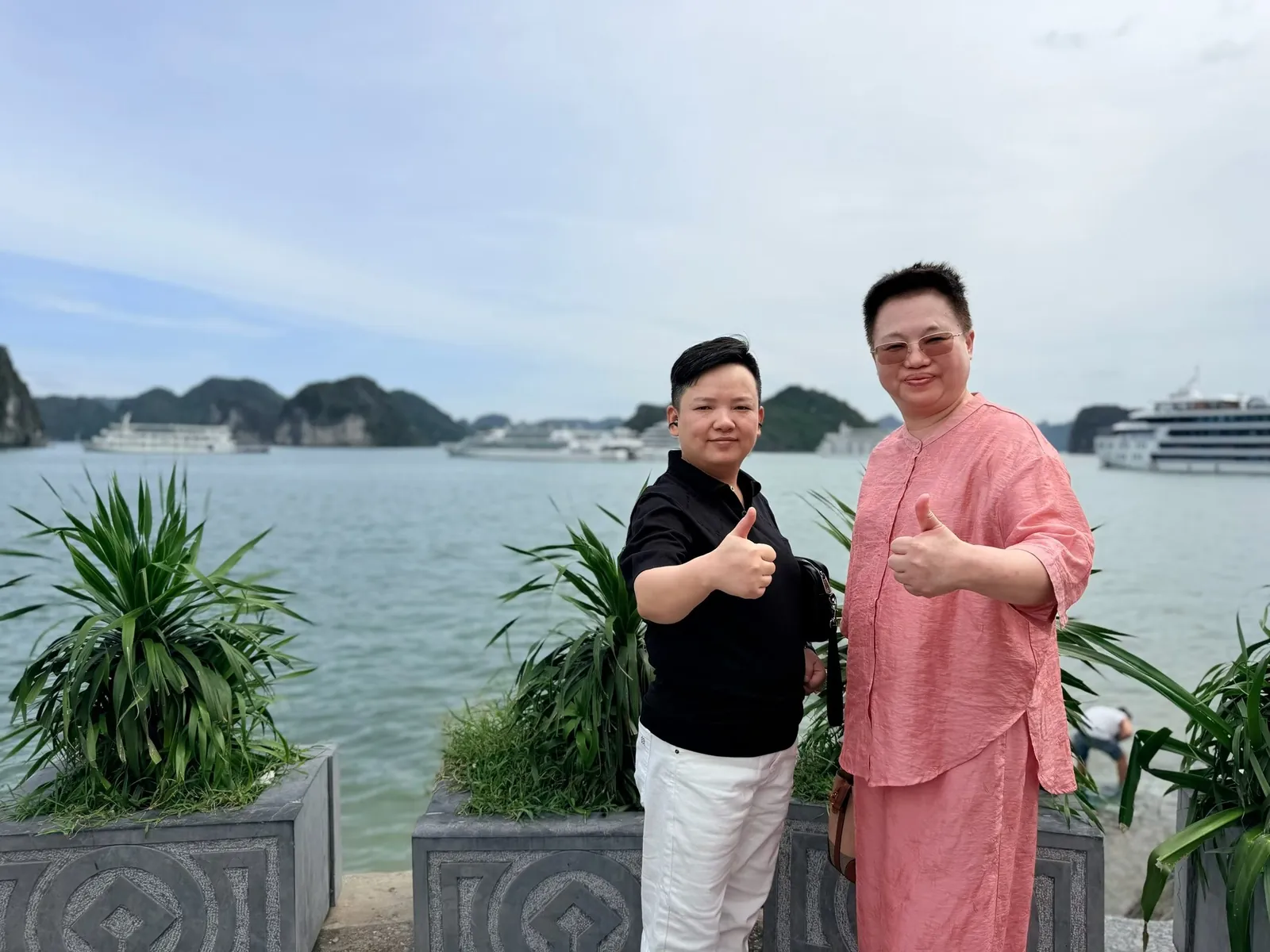 Two people giving a thumbs-up on the shore of Ha Long Bay, with the water, cruise ships and distant peaks behind them