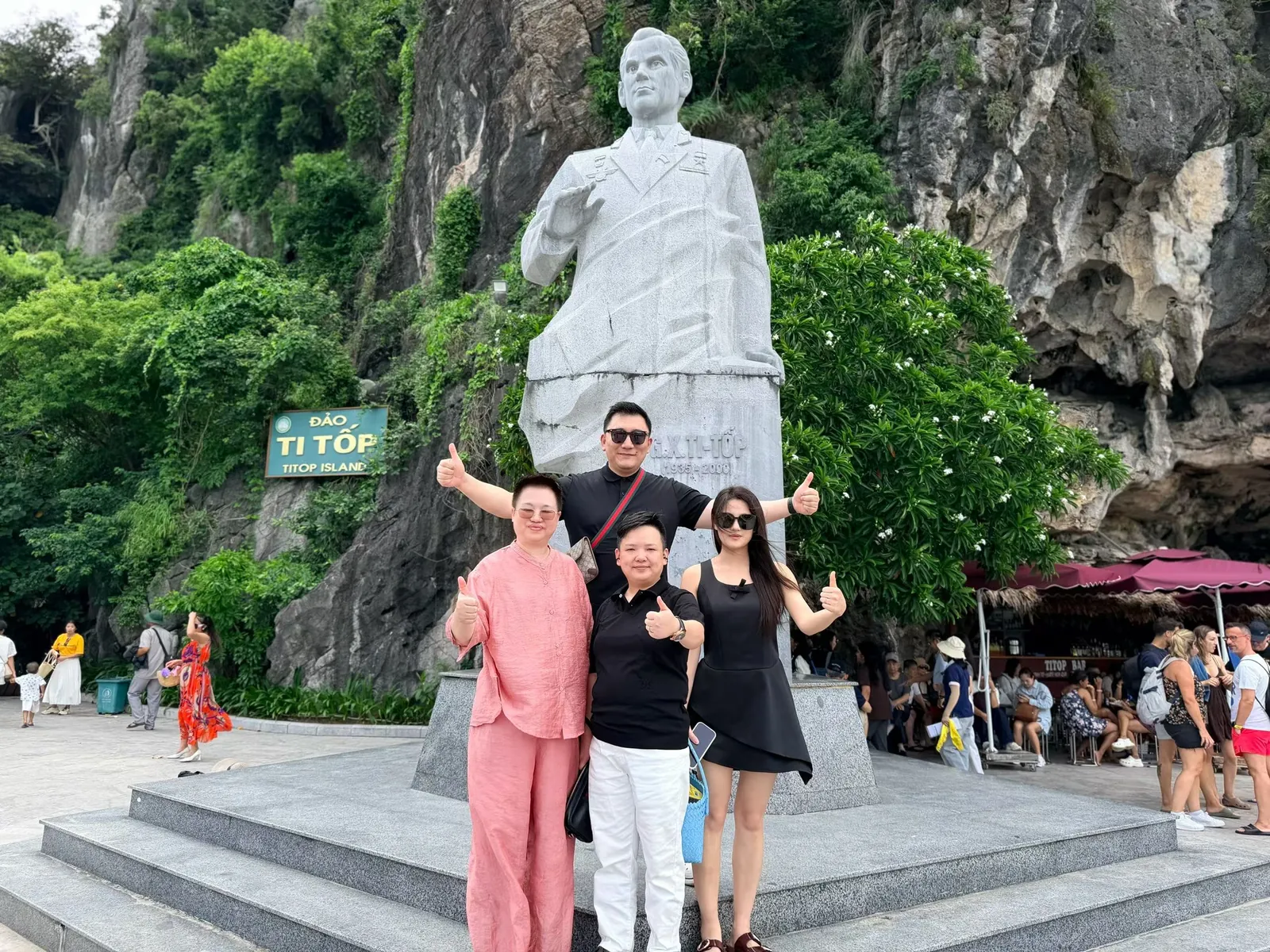 Four people giving a thumbs-up in front of the Titov statue on Titop Island, with a tree-covered limestone cliff behind them
