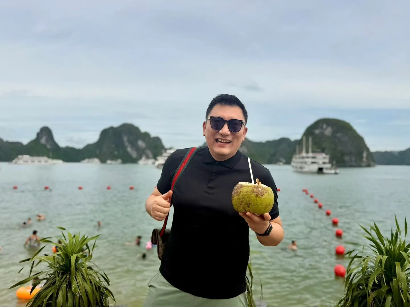 A man in sunglasses holding a coconut, with the beach, cruise boats and limestone islets of Ha Long Bay behind him